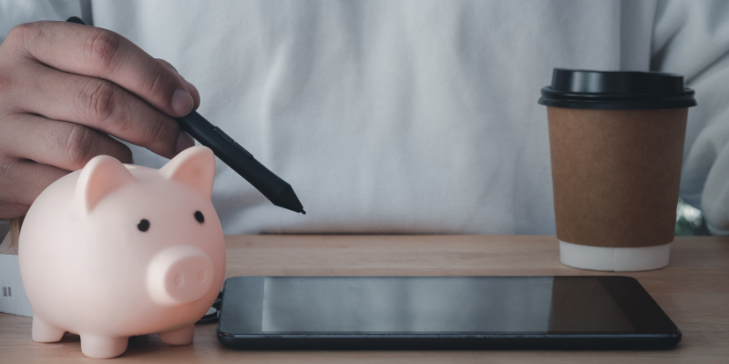 Close up of person working on tablet, with a take away coffee cup and a pink pig piggy bank next to the tablet.